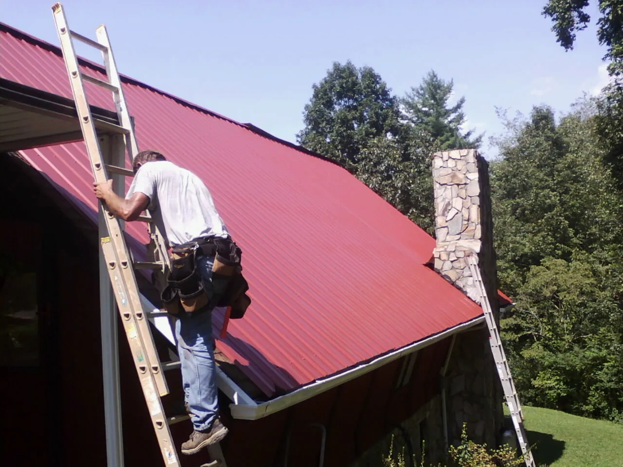 Roofer on a ladder beside a red metal roof and stone chimney — Soffit & Fascia Repair by Roofing Brothers in Long Valley